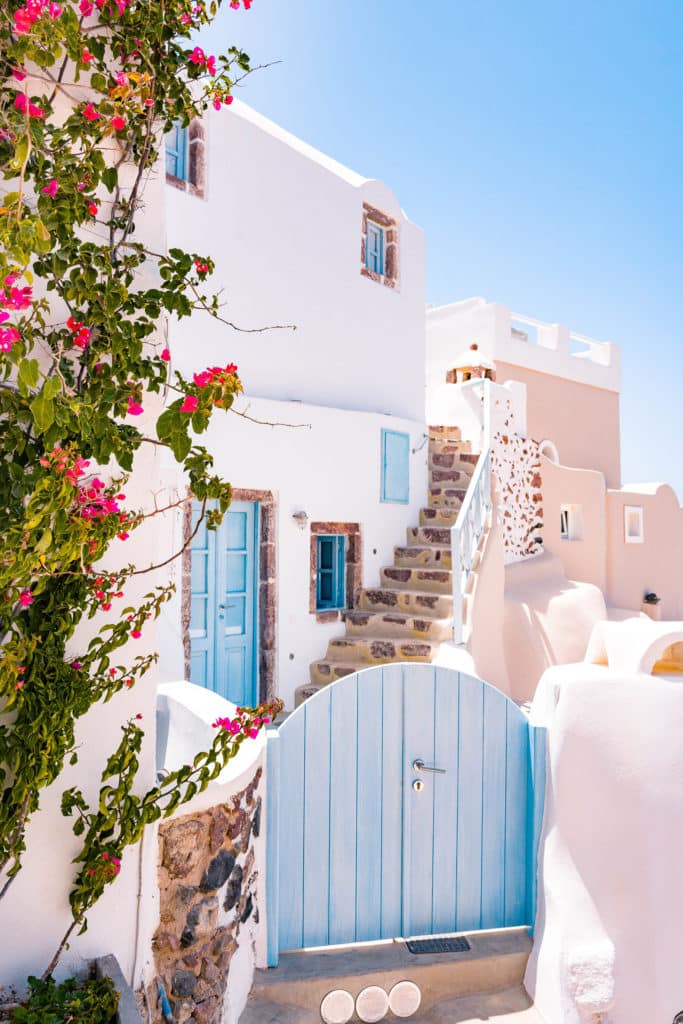 A quaint white villa in Greece with a blooming tree, blue fence, stone steps, during a bright and beautiful day.