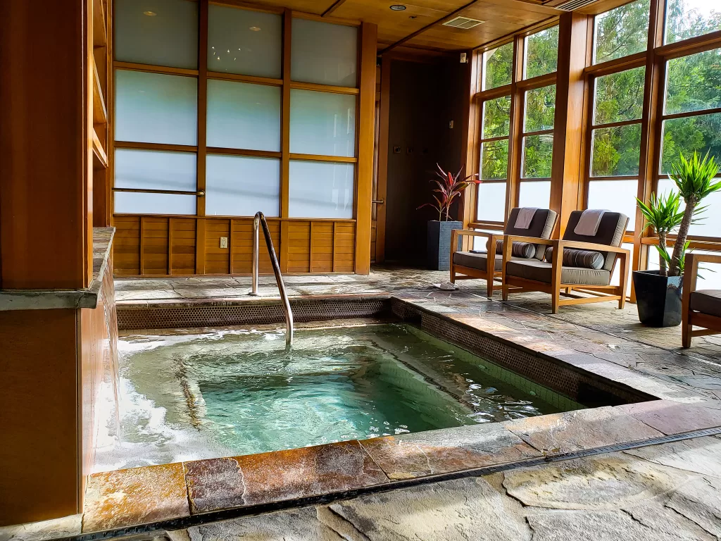 Salish Lodge and Spa mineral soaking pool with glass windows and lounge chairs. There is a view of evergreen trees outside and is a perfect location for a couple's getaway. 